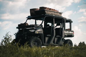 Front angled view of UTV featuring Swamp Ox hood rack, roof rack, and bed racks in grassy outdoor field. Black textured powder-coated racks all carrying outdoor gear.