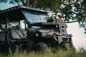 Angled front view of UTV featuring Swamp Ox hood rack, roof rack, and bed racks in grassy outdoor field. Black textured powder-coated racks all carrying outdoor gear.